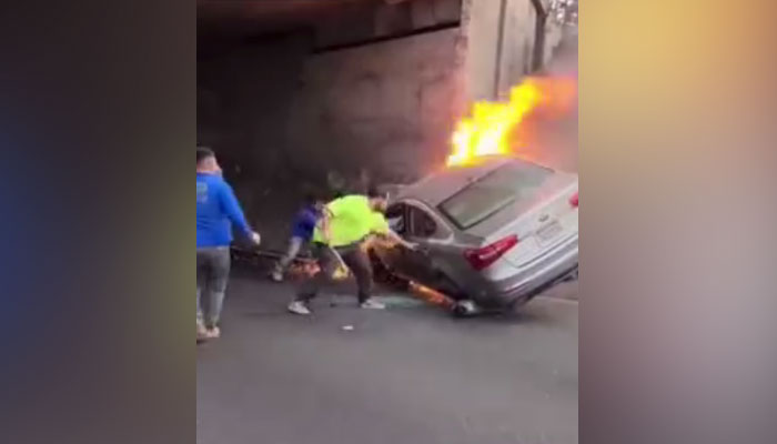 Un grupo de personas trata de rescatar a dos personas de un coche que se estrelló contra un pilar de hormigón y empezó a arder